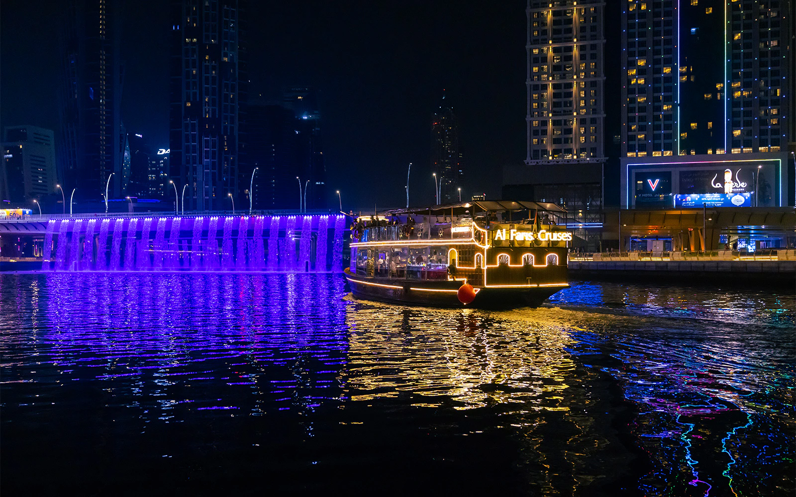 Dubai Canal cruise boat illuminated at night with city skyline and waterfall bridge.