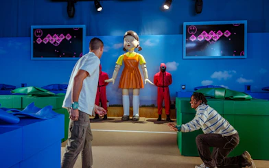 Participants in a Squid Game-themed play area with a large doll and guards in Dallas.