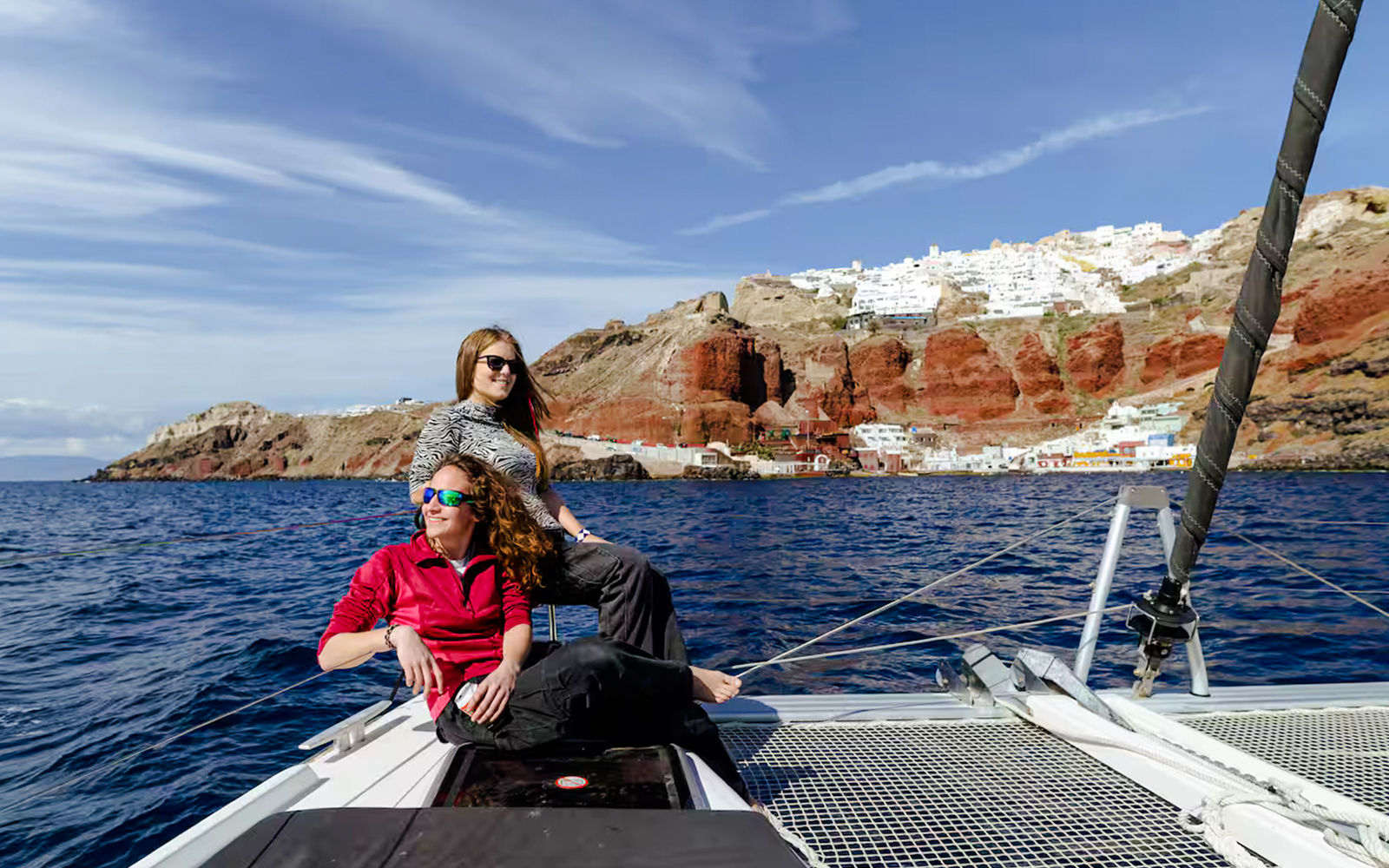 Guests relaxing on a catamaran with Santorini cliffs in the background during daytime cruise.