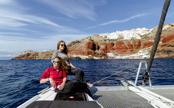 Guests relaxing on a catamaran with Santorini cliffs in the background during daytime cruise.