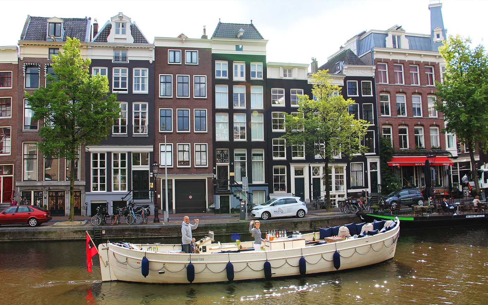 Canal boat with passengers enjoying drinks in front of Amsterdam's historic buildings.