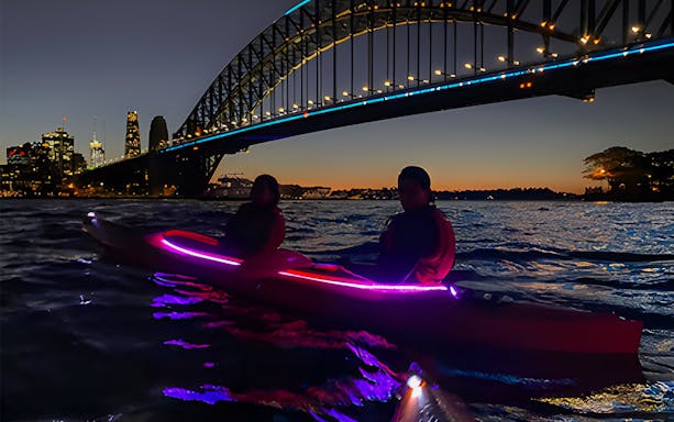 Group kayaking in LED-lit kayaks on Sydney Harbour at night with Luna Park and city skyline in view.