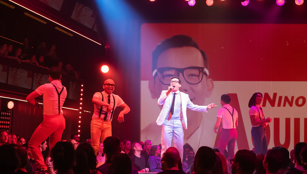 Performer singing on stage during "Here Lies Love" show with dancers in background.