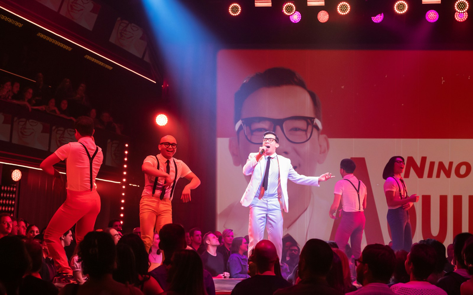 Performer singing on stage during "Here Lies Love" show with dancers in background.