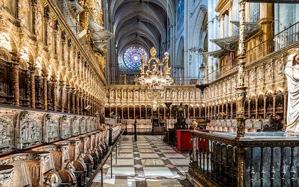 Interior of the Primate Cathedral of Saint Mary in Toledo, Spain, featuring ornate choir stalls and a rose window.