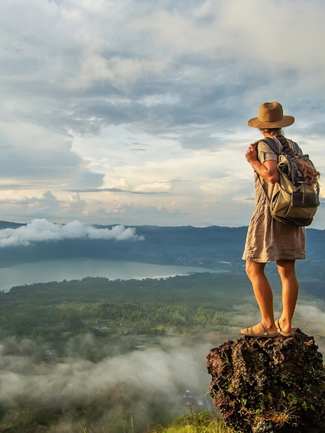 Woman standing on Mount Batur, Bali, enjoying sunrise view over clouds and landscape.