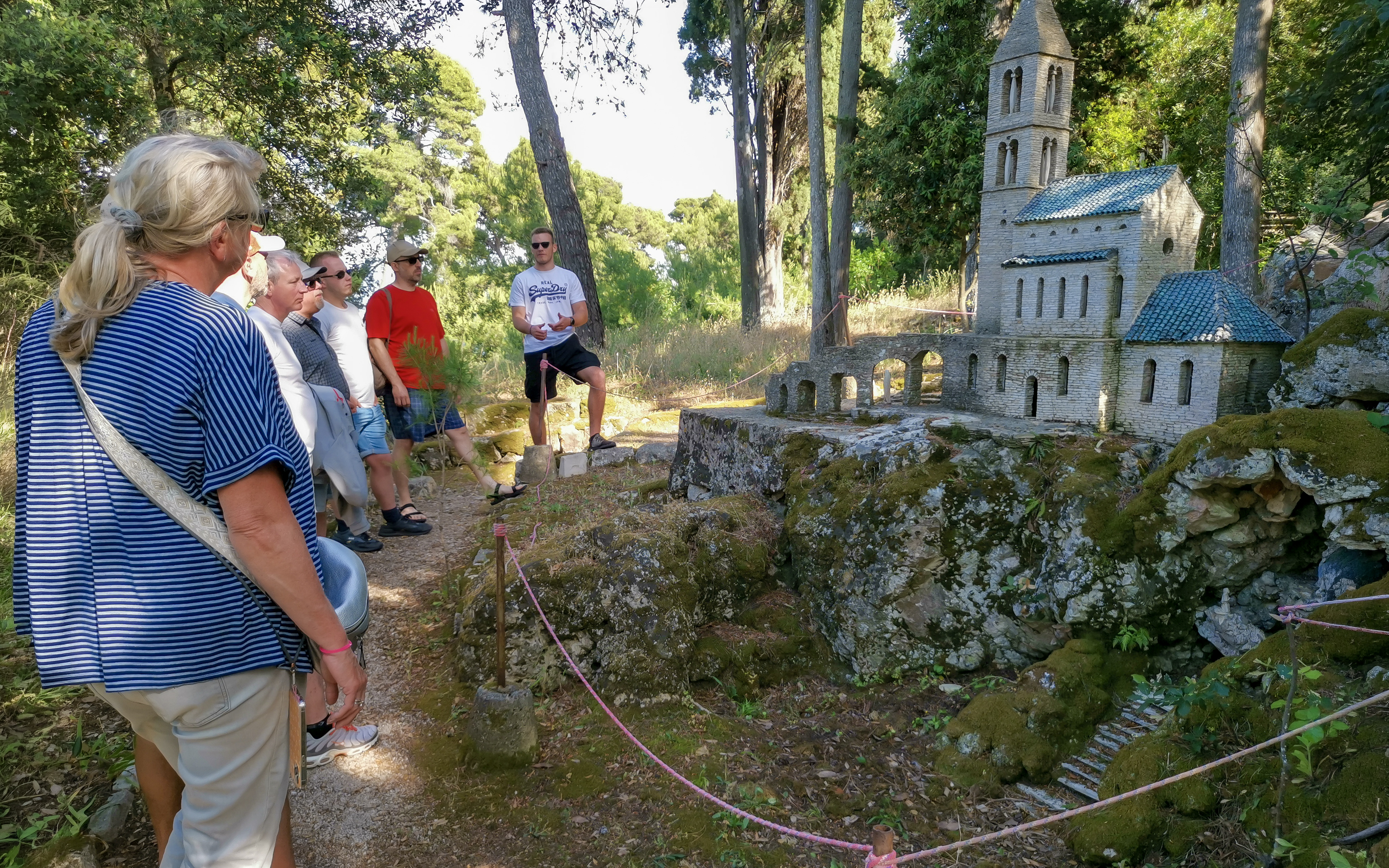 Tour group exploring a miniature stone church model in a forest near Zadar.
