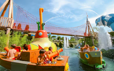 Children enjoying water ride at Oltremare park with colorful boats and bridge in background.