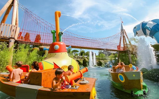 Children enjoying water ride at Oltremare park with colorful boats and bridge in background.