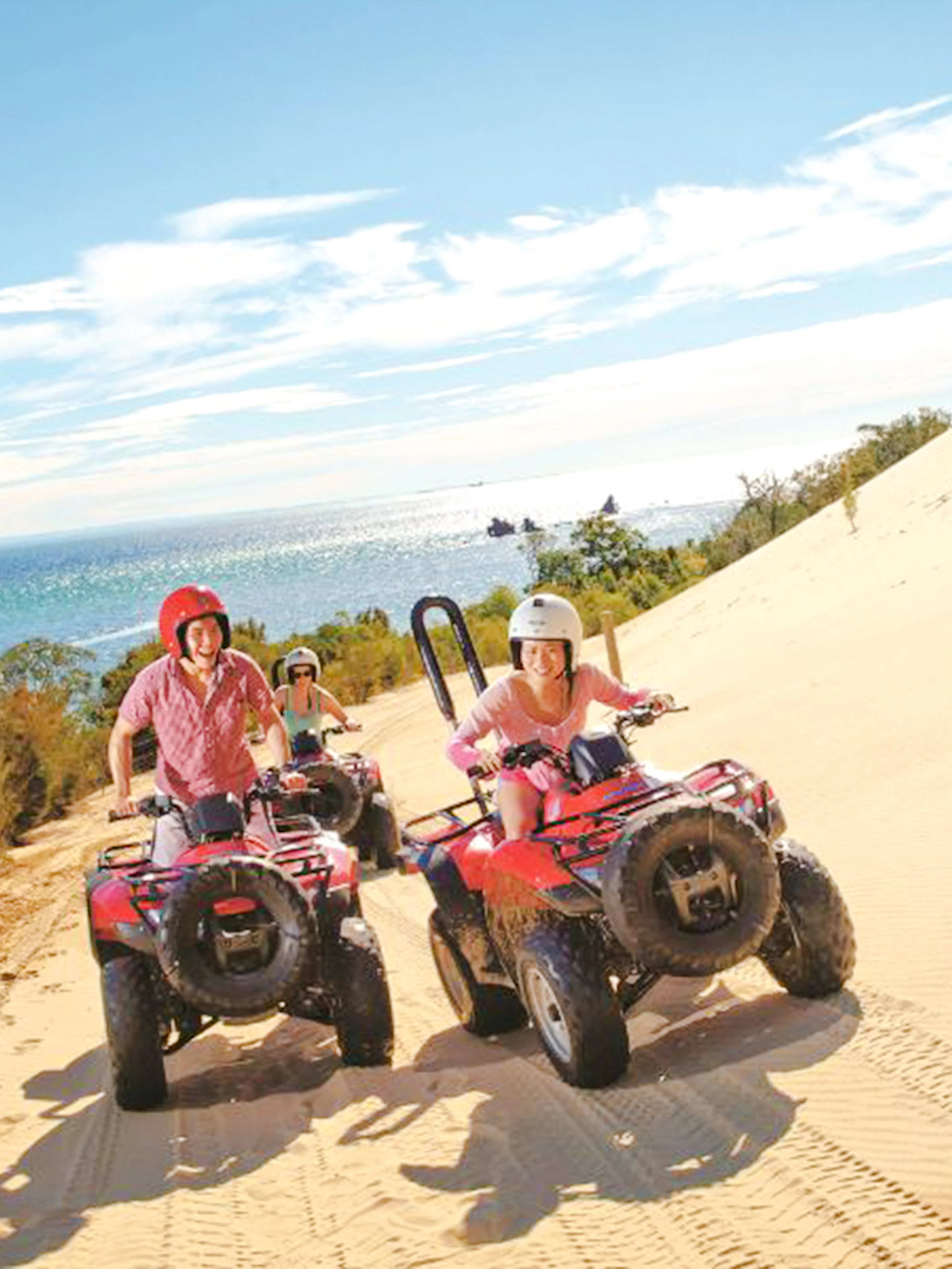 Riders on red quad bikes traverse a sand dune with ocean views on Moreton Island.