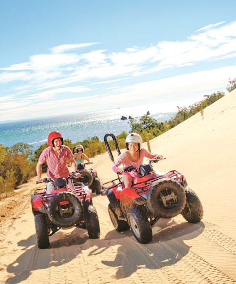 Riders on red quad bikes traverse a sand dune with ocean views on Moreton Island.