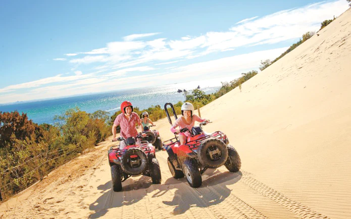 Riders on red quad bikes traverse a sand dune with ocean views on Moreton Island.