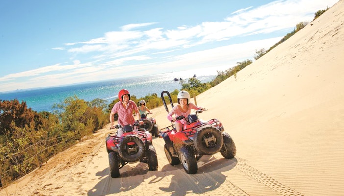 Riders on red quad bikes traverse a sand dune with ocean views on Moreton Island.