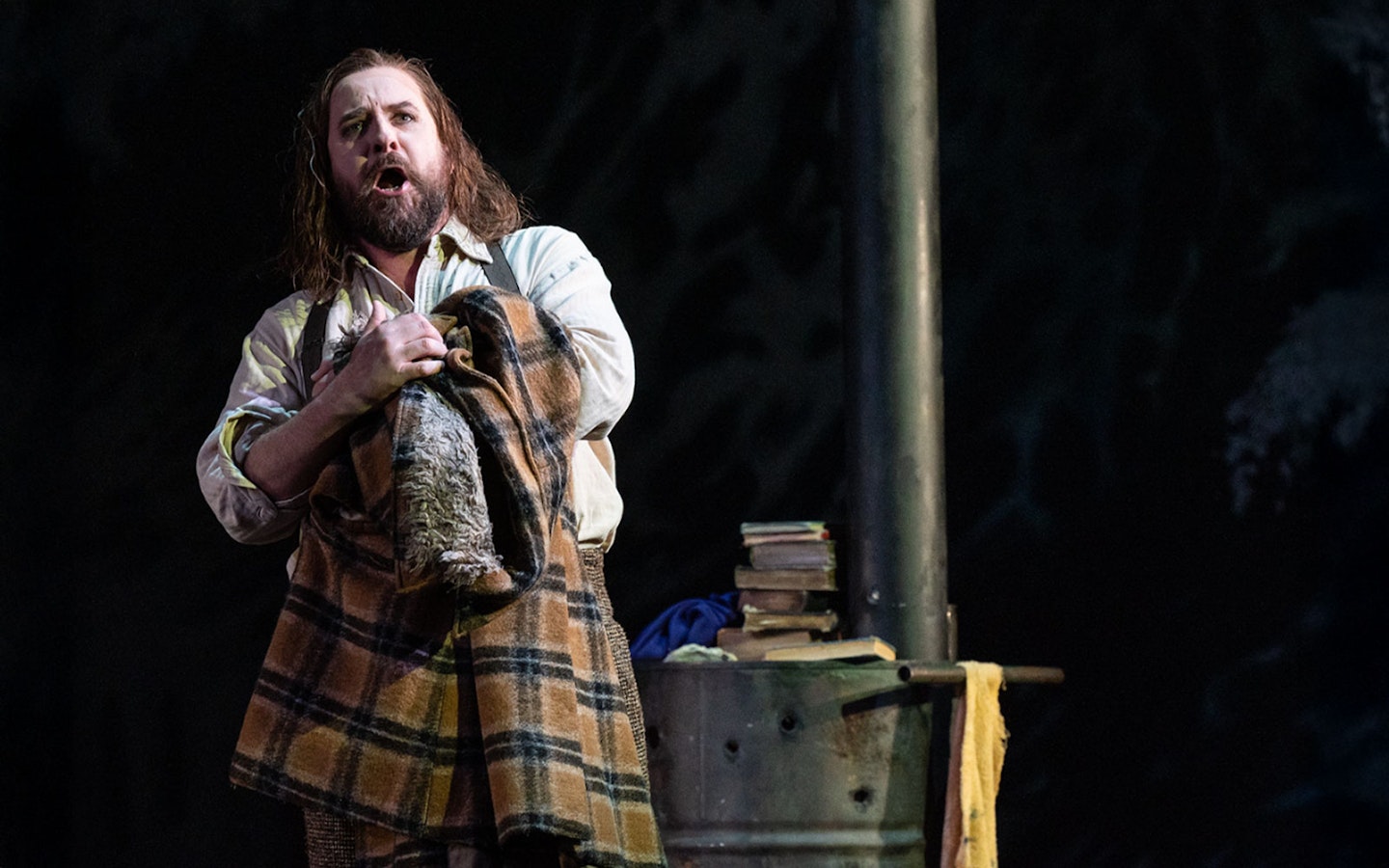 Opera performer in a scene from La Bohème, holding a plaid blanket on stage.