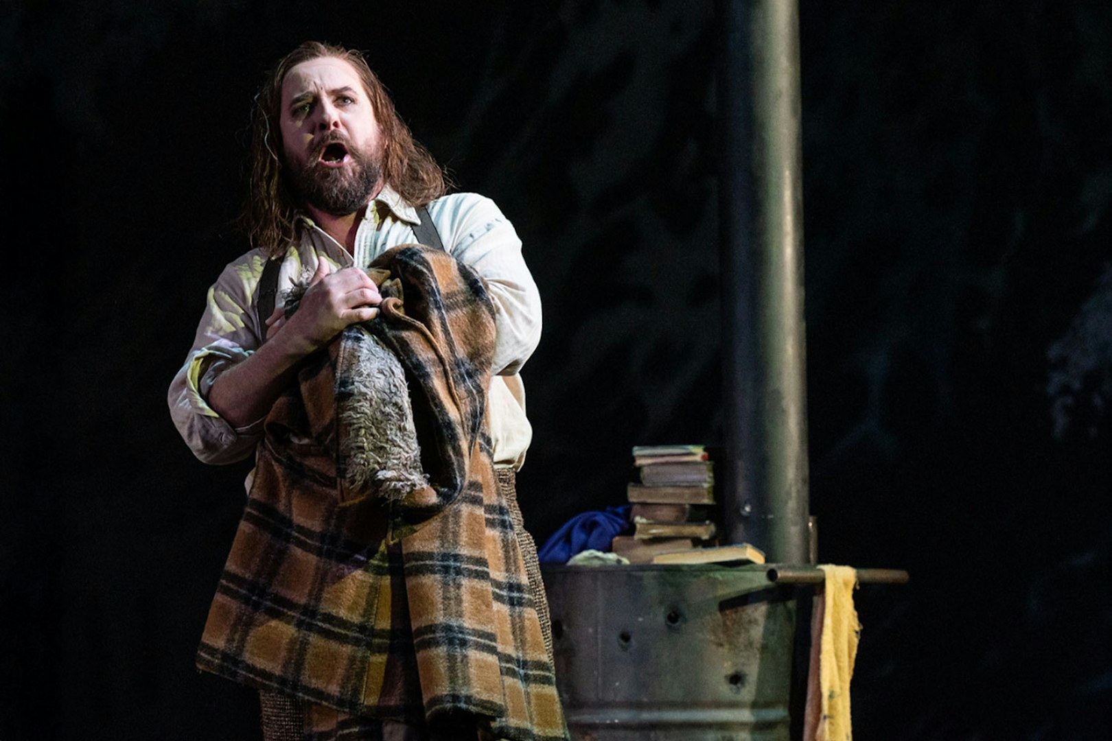 Opera performer in a scene from La Bohème, holding a plaid blanket on stage.
