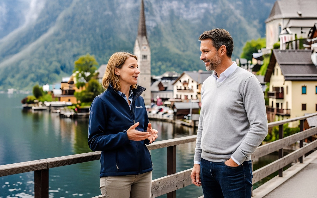 Tour guide discussing with a visitor on a bridge in Hallstatt, Austria, with lake and church in background.