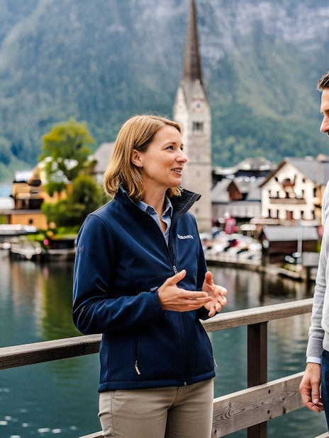 Tour guide discussing with a visitor on a bridge in Hallstatt, Austria, with lake and church in background.
