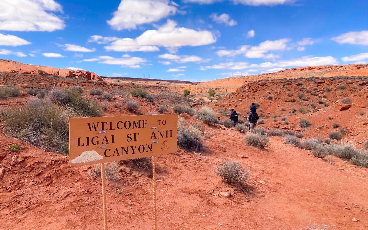Welcome sign at Ligai Si Anii Canyon with horseback riders on red desert trail.