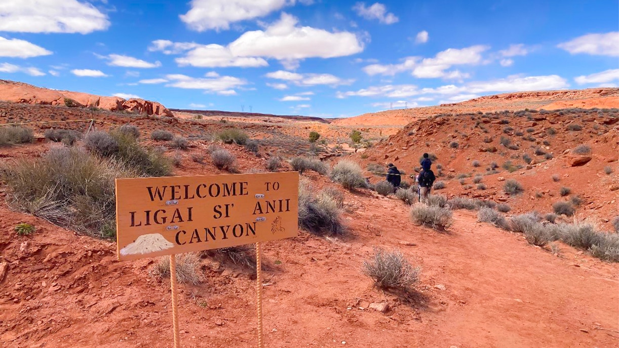 Welcome sign at Ligai Si Anii Canyon with horseback riders on red desert trail.