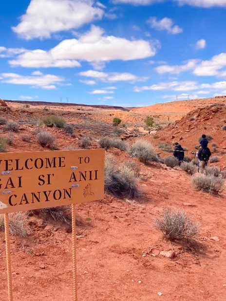 Welcome sign at Ligai Si Anii Canyon with horseback riders on red desert trail.