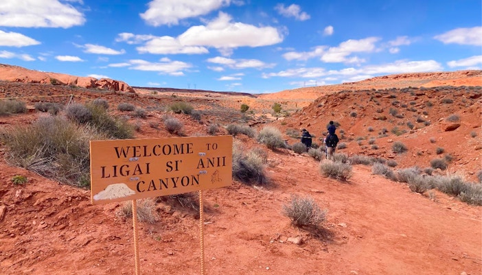 Welcome sign at Ligai Si Anii Canyon with horseback riders on red desert trail.