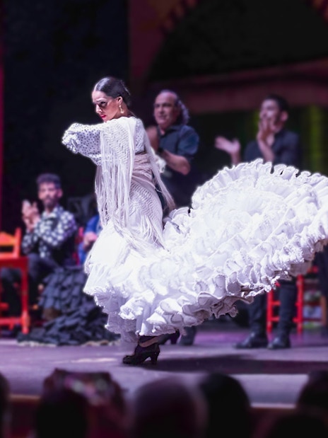 Flamenco dancer in white dress performing on stage with musicians in background.