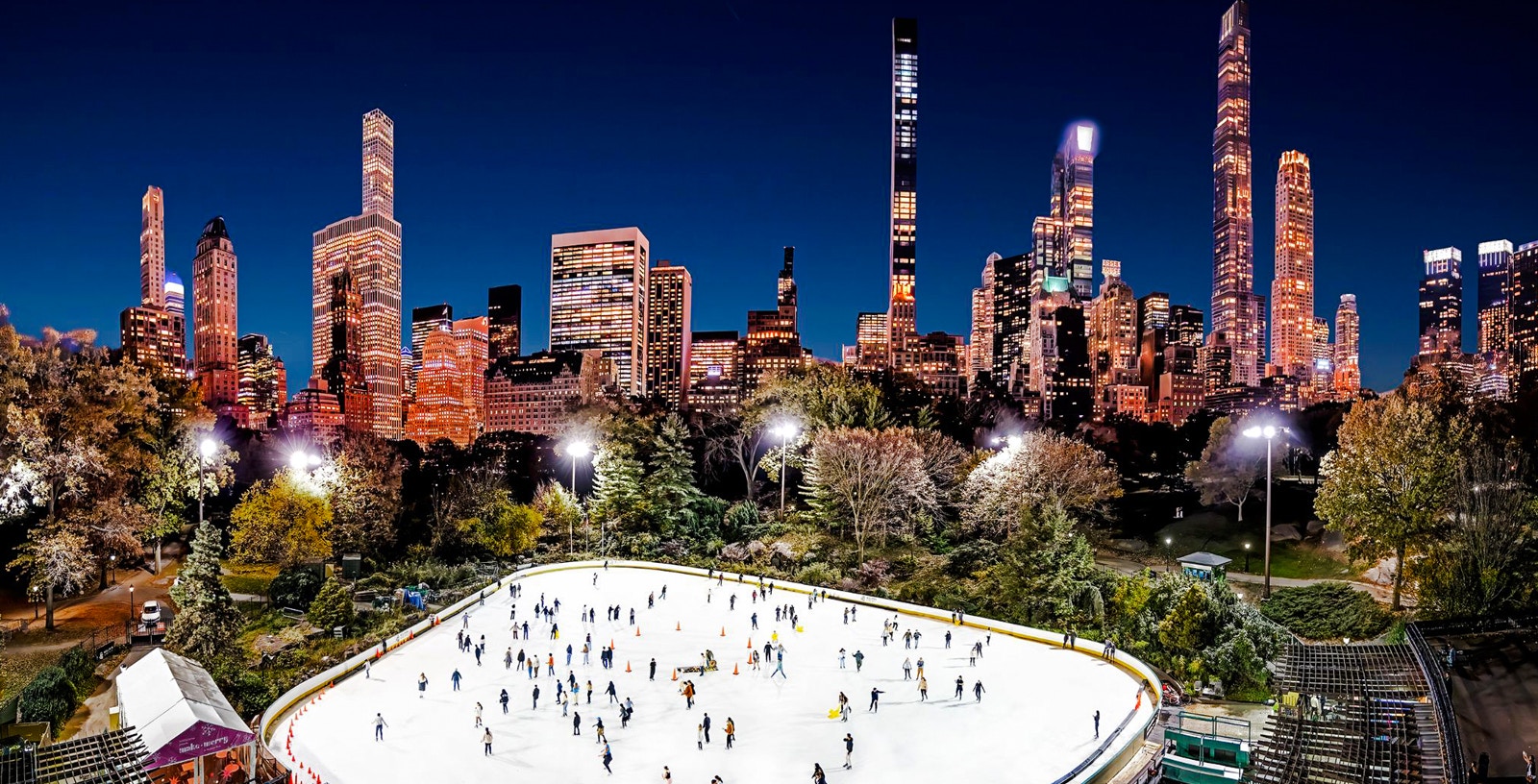 Wollman Ice Rink in Central Park with skaters and New York City skyline at night.