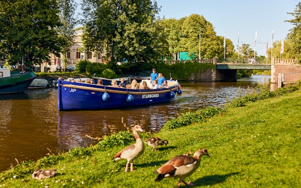 Amsterdam canal cruise boat with passengers enjoying drinks, passing under a bridge.