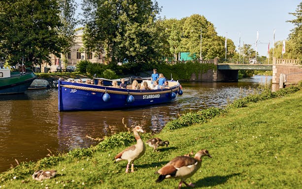 Amsterdam canal cruise boat with passengers enjoying drinks, passing under a bridge.