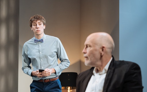 Two men in a room, one holding a phone, related to Bitter Wheat play.