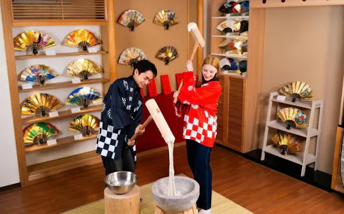 Couple participating in mochi pounding at Tokyo experience with decorative fans in background.
