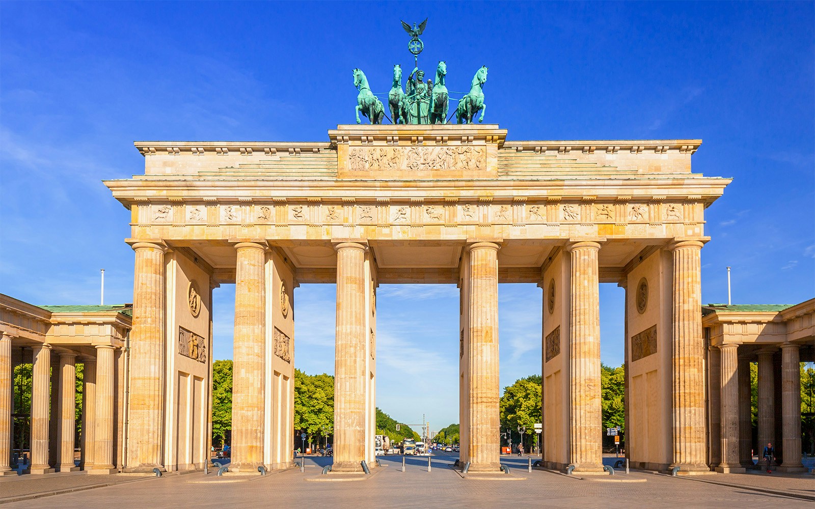 Das Brandenburger Tor in Berlin bei klarem, sonnigem Himmel, umgeben von Bäumen und Straßen.