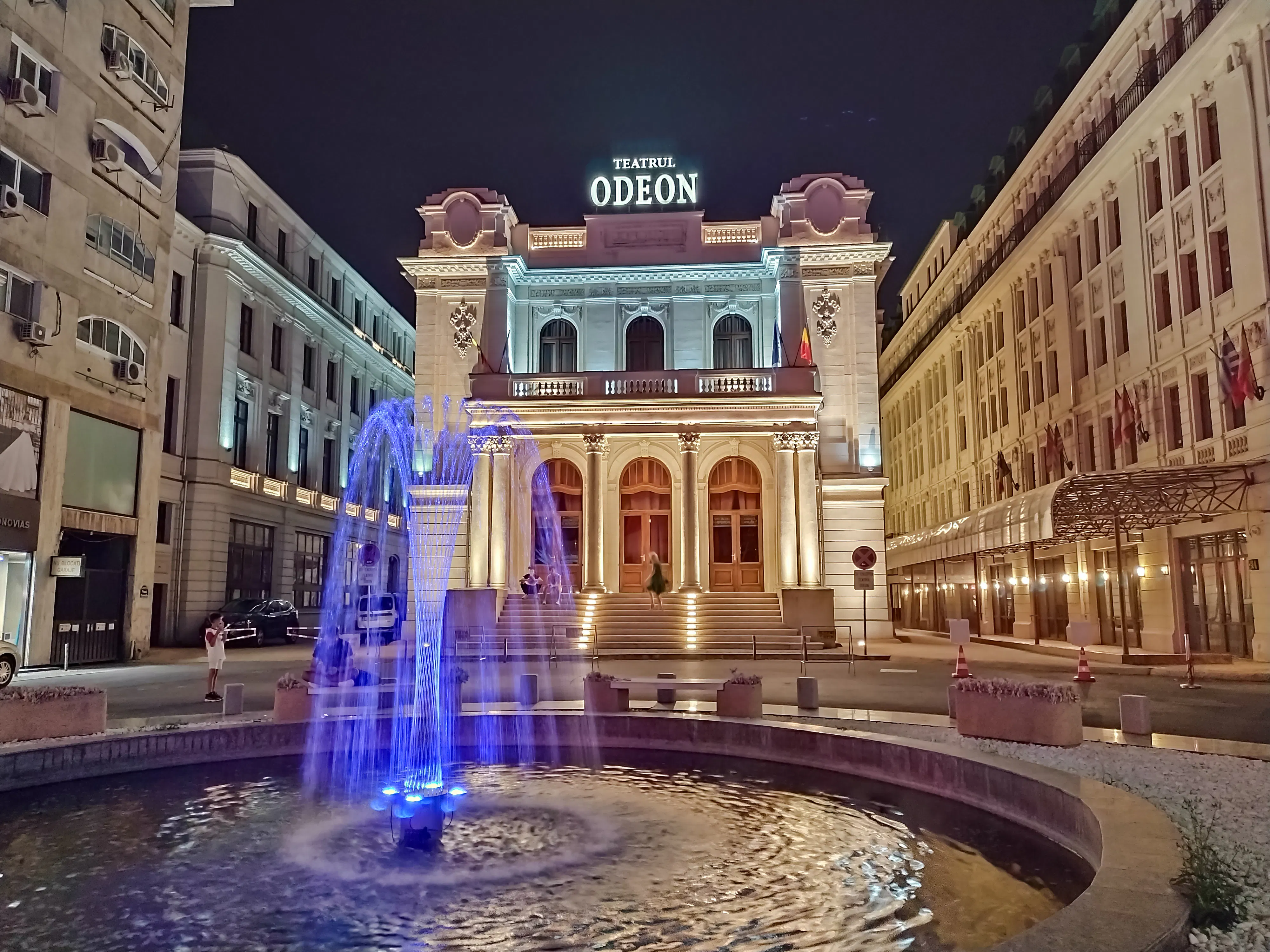 Odeon Theatre illuminated at night with a blue-lit fountain in Bucharest.