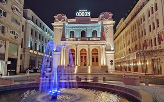 Odeon Theatre illuminated at night with a blue-lit fountain in Bucharest.