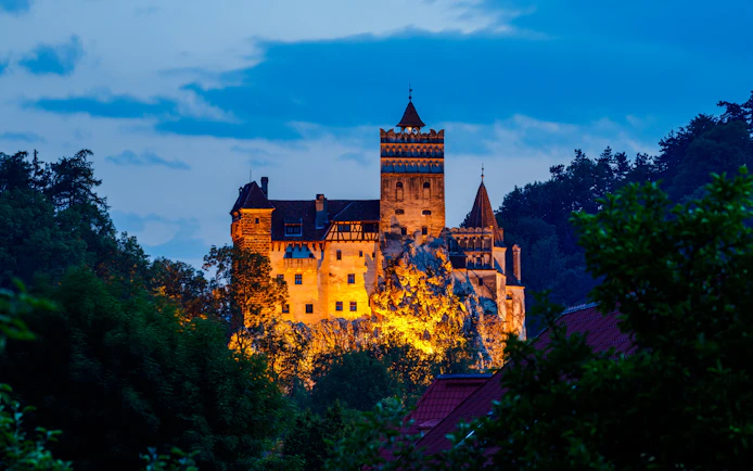 Bran Castle illuminated at dusk, surrounded by trees, Romania.