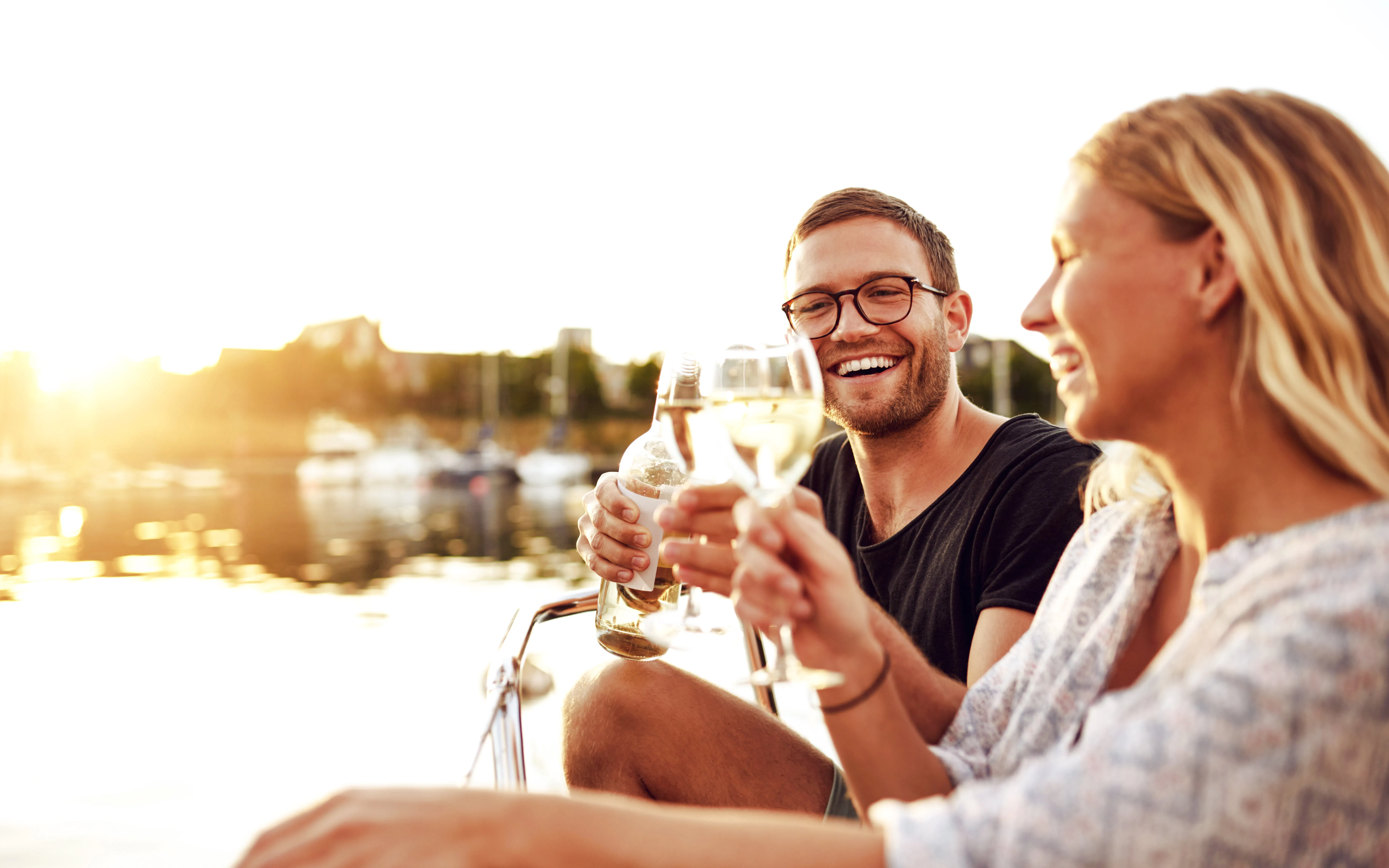 Couple toasting wine glasses on a boat at sunset, marina in the background.