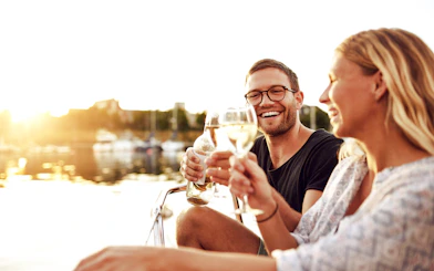 Couple toasting wine glasses on a boat at sunset, marina in the background.