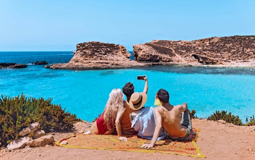 Friends sitting on a cliff, taking a selfie overlooking the Blue Lagoon, Comino Island.