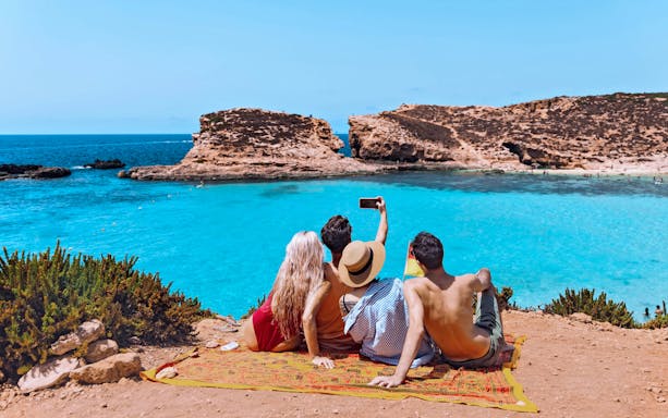 Friends sitting on a cliff, taking a selfie overlooking the Blue Lagoon, Comino Island.