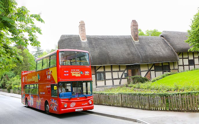Red double-decker HOHO tour bus in Stratford passing a thatched cottage.
