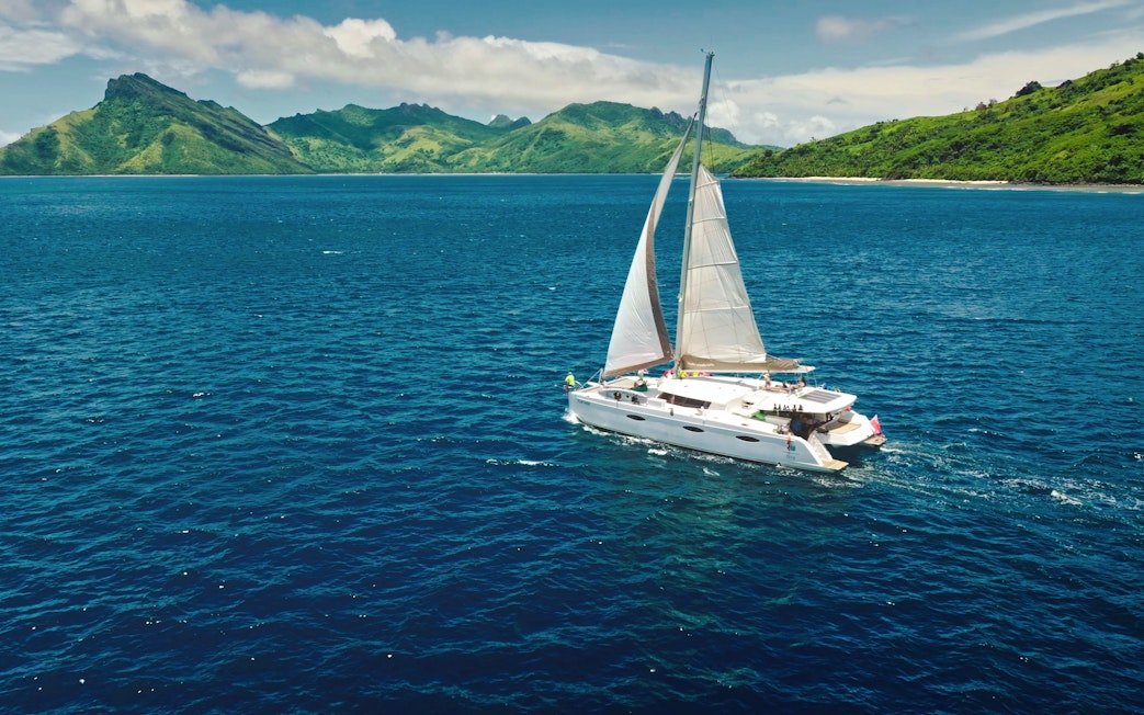 Sailing catamaran near Kuata Island, Fiji, with lush green hills in the background.