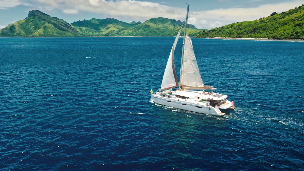 Sailing catamaran near Kuata Island, Fiji, with lush green hills in the background.
