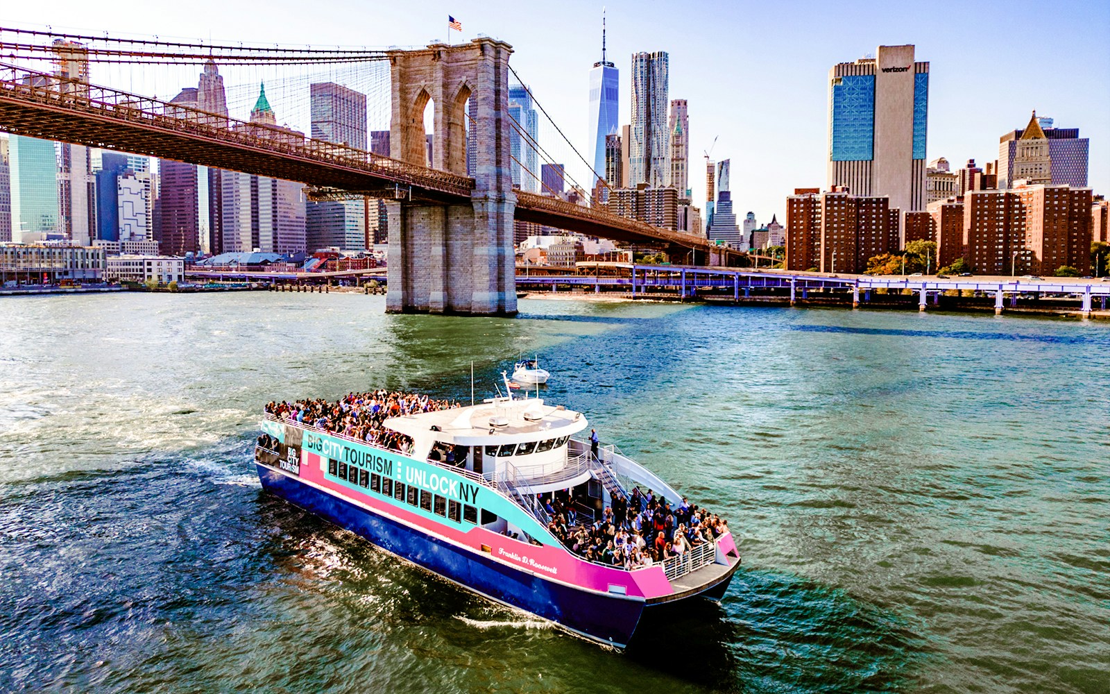 Cruise boat near Brooklyn Bridge, New York City skyline in background.