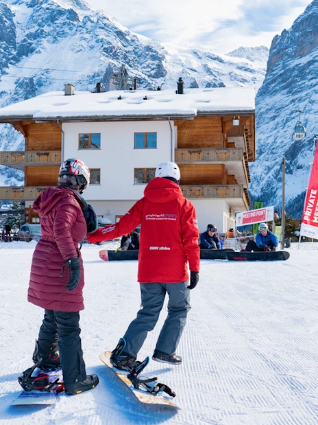 Beginner snowboarders at Grindelwald with snowy mountains and chalet in background.