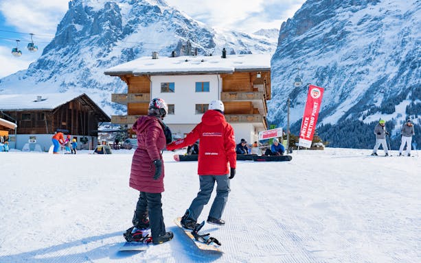 Beginner snowboarders at Grindelwald with snowy mountains and chalet in background.