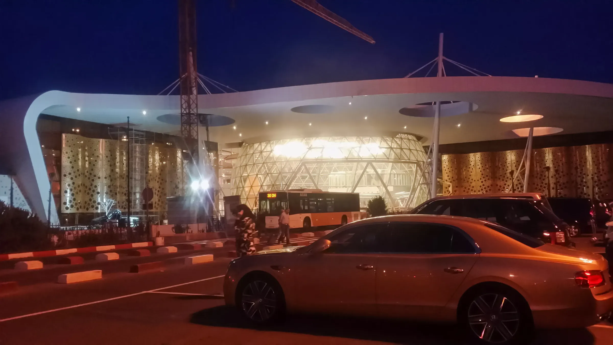 Marrakech Airport at night with parked cars and a bus in front of the terminal.