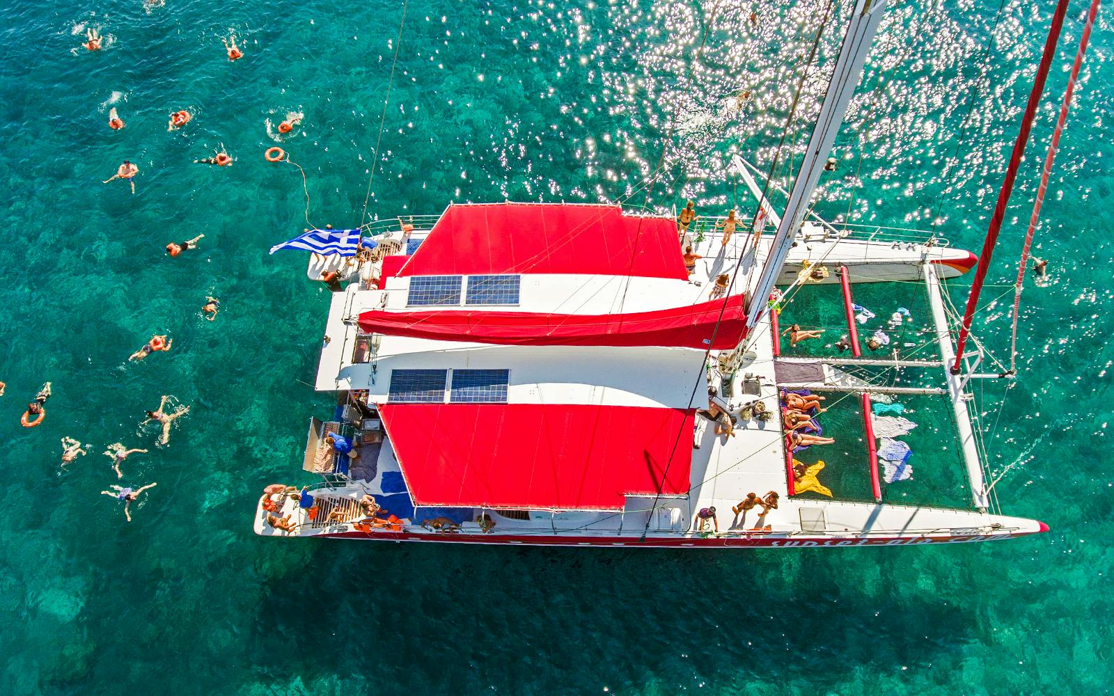 Catamaran with red sails on clear blue water, people swimming nearby, Greece flag visible.
