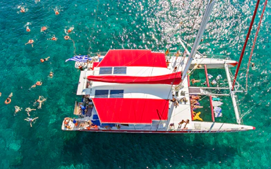 Catamaran with red sails on clear blue water, people swimming nearby, Greece flag visible.