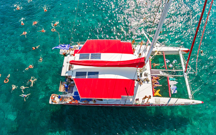 Catamaran with red sails on clear blue water, people swimming nearby, Greece flag visible.