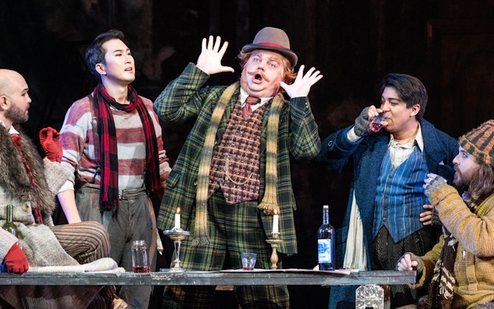 Opera performers in a lively scene from La Bohème, with expressive gestures and period costumes.
